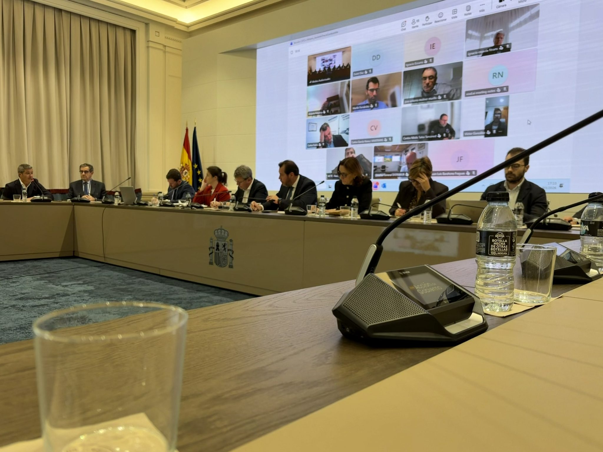 Reunión en el Ministerio de Transportes y Movilidad Sostenible: representantes de la Administración y de las organizaciones del transporte en torno a una mesa, con las banderas de España y de la Unión Europea y participantes conectados por videoconferencia en una gran pantalla