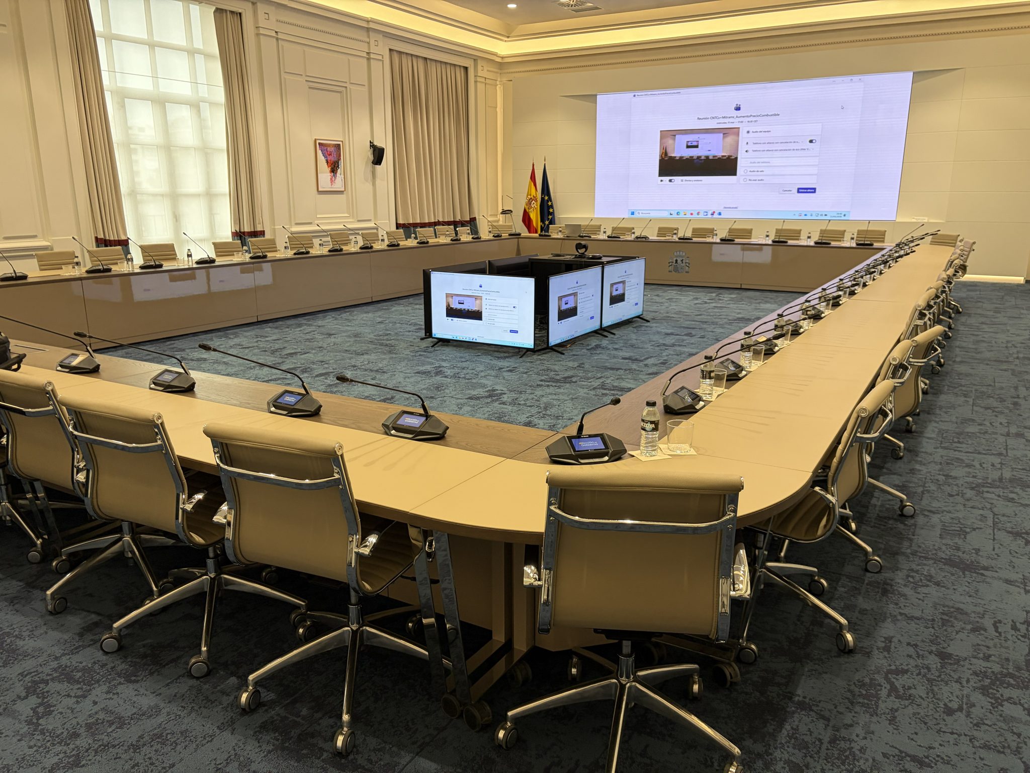 Sala de reuniones del Ministerio de Transportes y Movilidad Sostenible antes de la reunión, con una gran mesa en forma de U, micrófonos en cada puesto, pantallas y las banderas de España y de la Unión Europea